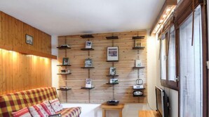 Un salon chaleureux avec un canapé à carreaux, des étagères murales en bois ornées de divers objets, une petite table en bois et un téléviseur posé sur un meuble.