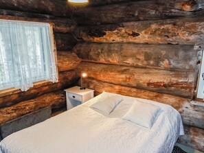 A wooden cabin room with a bed, a nightstand, and a window with lace curtains. - Villa mitja by Interhome (Posio)