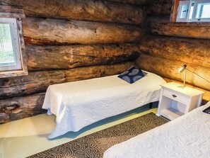 A log cabin room with two beds, a nightstand, a lamp, and a window with blinds. - Villa mitja by Interhome (Posio)