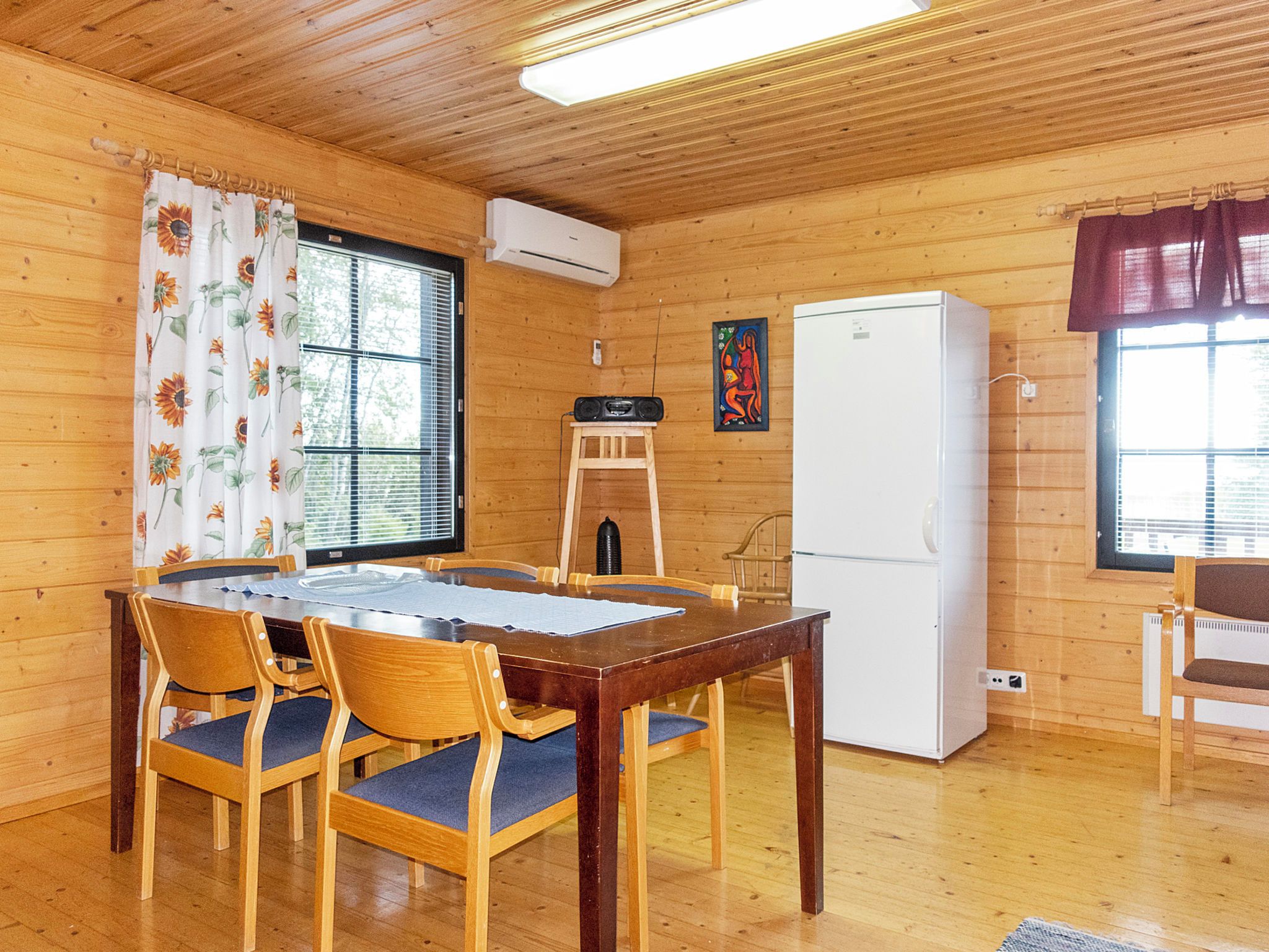 En stuga med träpanel på insidan, inredd med ett matbord, stolar, ett kylskåp och ett fönster med persienner.