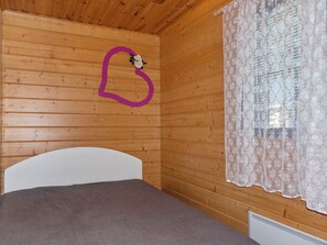 A wooden room with a bed, a window with floral curtains, and a purple heart-shaped decoration on the wall.