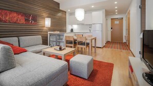 Un salon moderne avec un canapé gris, une table basse et un tapis rouge. On y trouve également un coin cuisine avec des meubles blancs et un réfrigérateur.