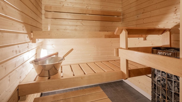 Un sauna avec des murs et des bancs en bois, un seau en métal et un récipient rempli de pierres portant l’inscription « HARVIA ».