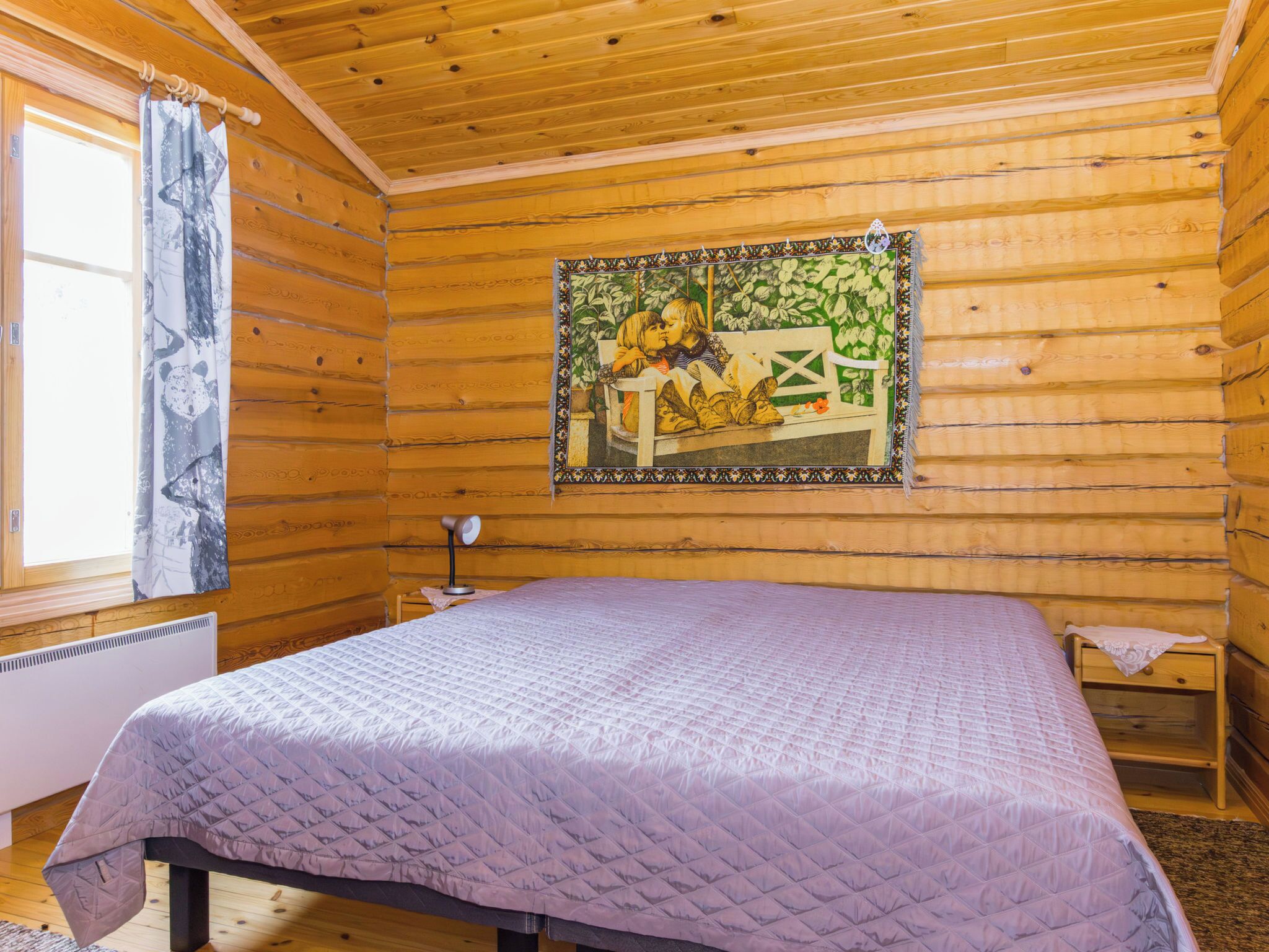 Ein Zimmer einer Holzhütte mit einem Bett, einem Vorhangfenster, einem Heizkörper und einer Wandstickerei.