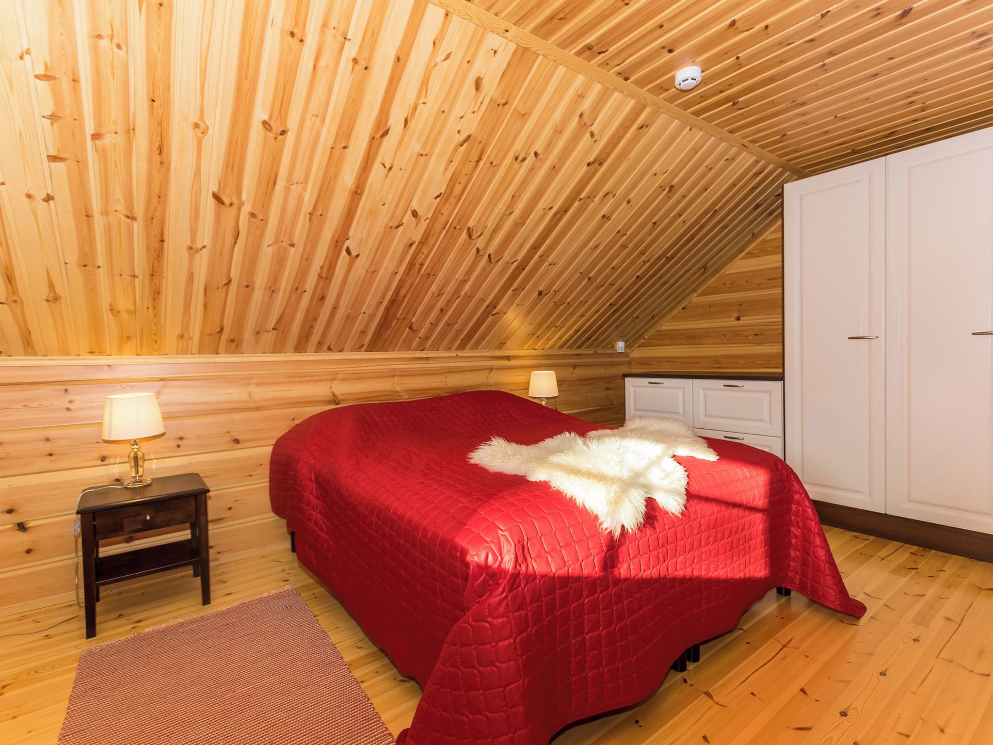 Une pièce avec un plafond en bois incliné, un lit recouvert d’une courtepointe rouge, un plaid en fausse fourrure blanche, une petite table de chevet avec une lampe et une armoire.