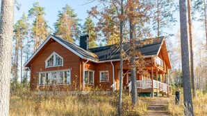 Een houten chalet met een veranda en schoorsteen, omgeven door hoge bomen en een pad dat naar de ingang leidt.