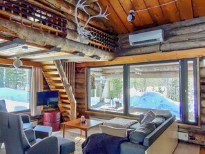 Interior de cabana de tronco com sofá, cadeiras, mesa, televisão e grandes janelas com vista para uma paisagem nevada.