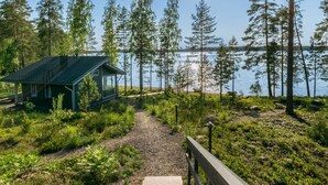 A cabin by a lake with a clear path leading to it, surrounded by tall trees and greenery.