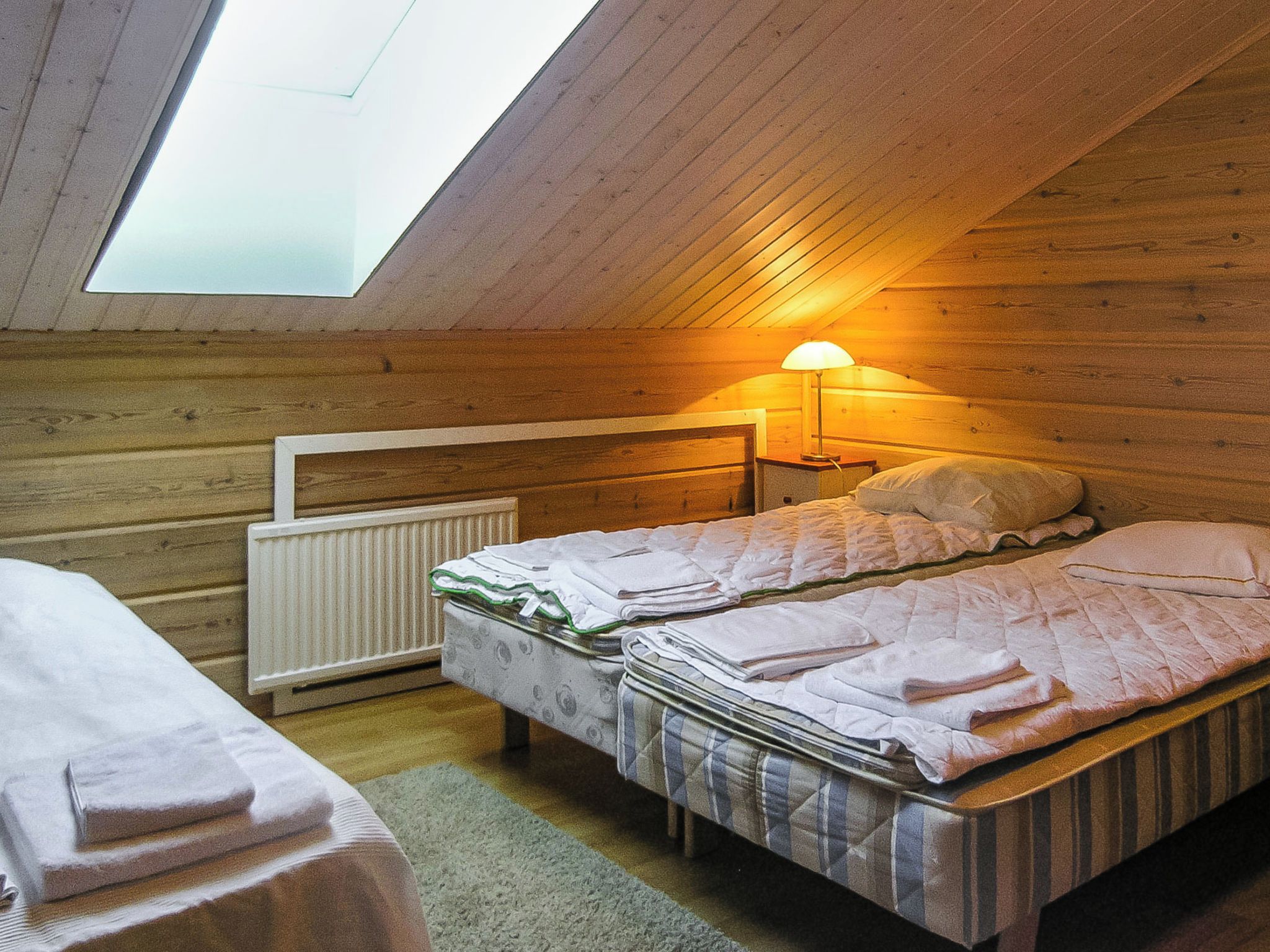 A small, cozy room with a sloped ceiling and a skylight, featuring a bed with a quilted blanket and pillows, a bedside table with a lamp, and a radiator.