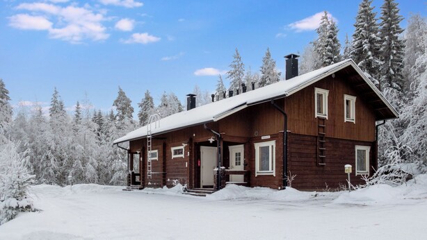 A wooden cabin with a snow-covered roof, surrounded by snow-laden trees, under a clear blue sky.