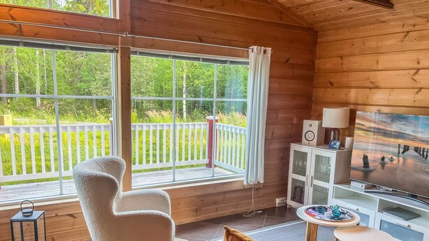 Un interior de cabaña acogedor con un ventanal grande, un sillón cómodo, un sofá, un televisor y una mesita.