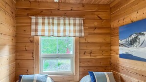 Habitación tipo cabaña con dos camas, una ventana con cortina a cuadros y un cuadro en la pared.