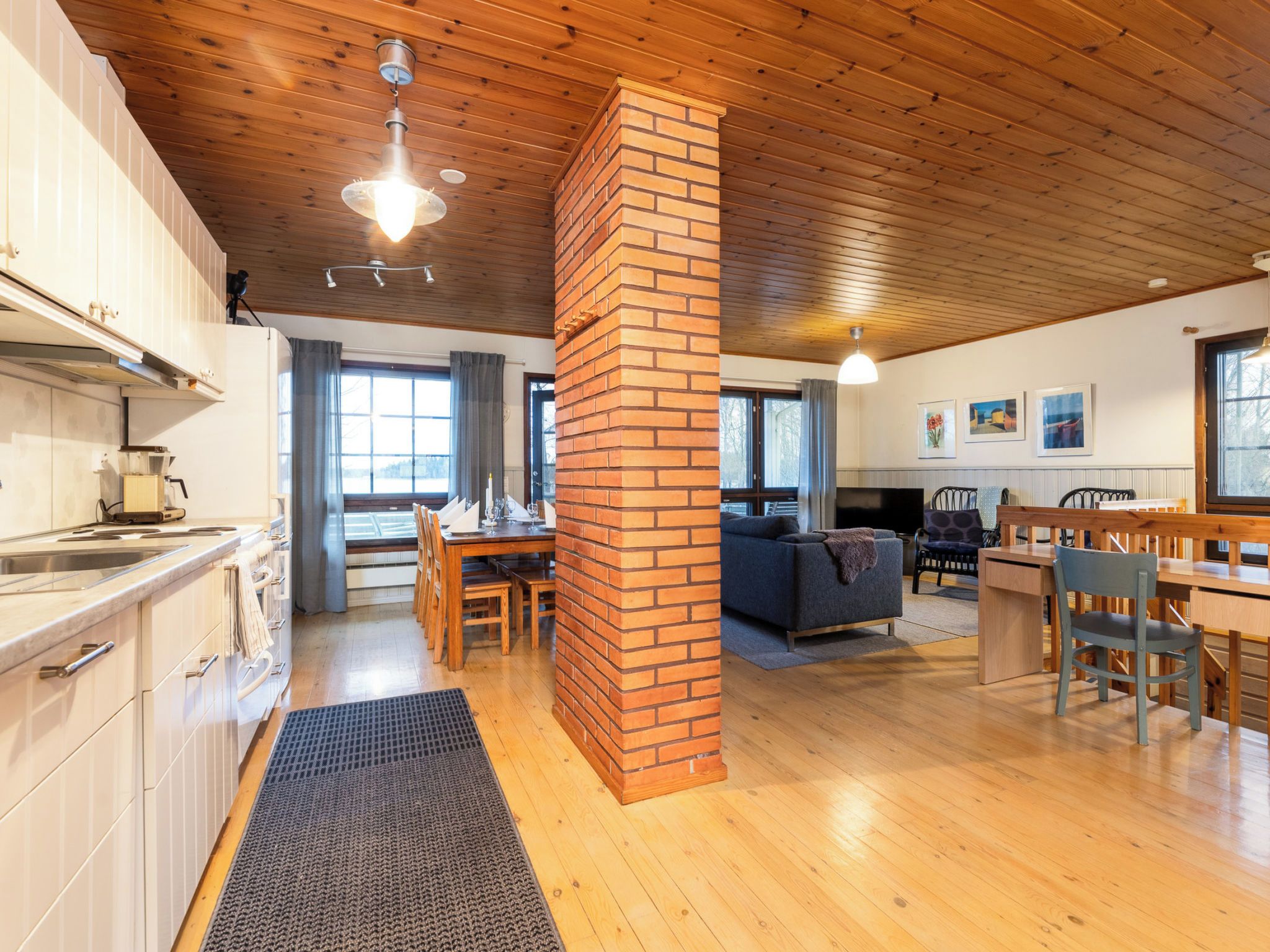 A spacious living area with a wooden ceiling, a brick column, a dining table with chairs, a sofa, and a TV.