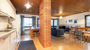 A spacious living area with a wooden ceiling, a brick column, a dining table with chairs, a sofa, and a TV.