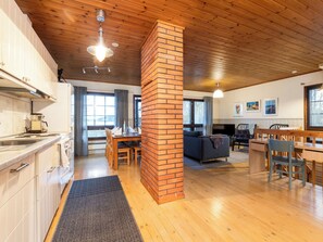 Een ruime woonkamer met houten plafond, een bakstenen zuil, een eettafel met stoelen, een bank en een televisie.
