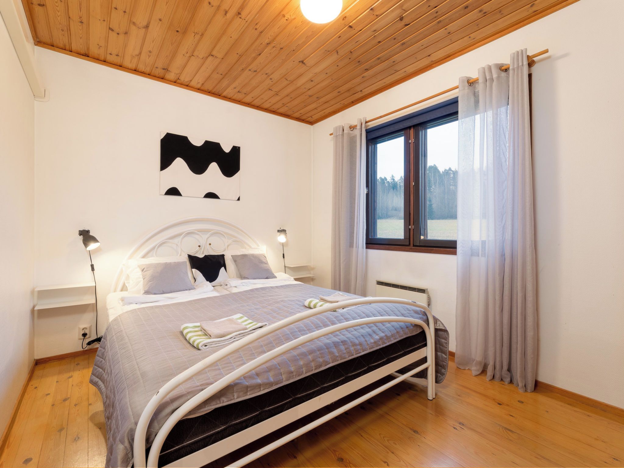 Un dormitorio con techo de madera, una cama con cabecera blanca, dos lámparas de noche, una ventana con cortinas y un radiador.
