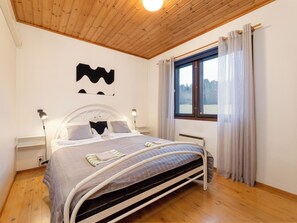 Un dormitorio con techo de madera, una cama con cabecera blanca, dos lámparas de noche, una ventana con cortinas y un radiador.