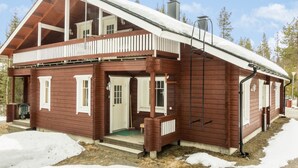 A two-story wooden cabin with a balcony, a chimney, and a snow-covered roof.