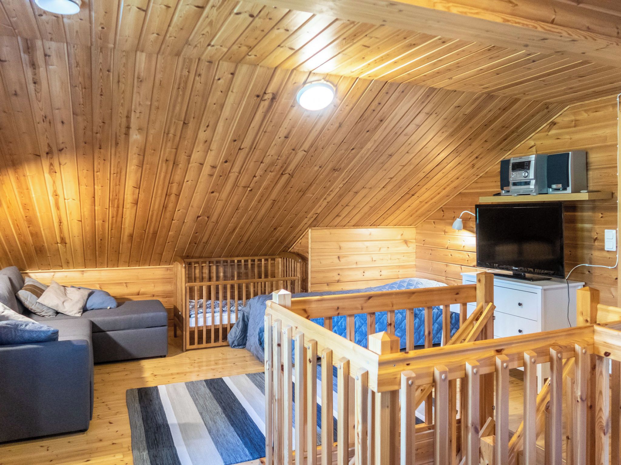 A cozy attic bedroom with a sloped ceiling, a bed with a blue quilt, a grey sofa, a small desk with a television, and a wooden crib.