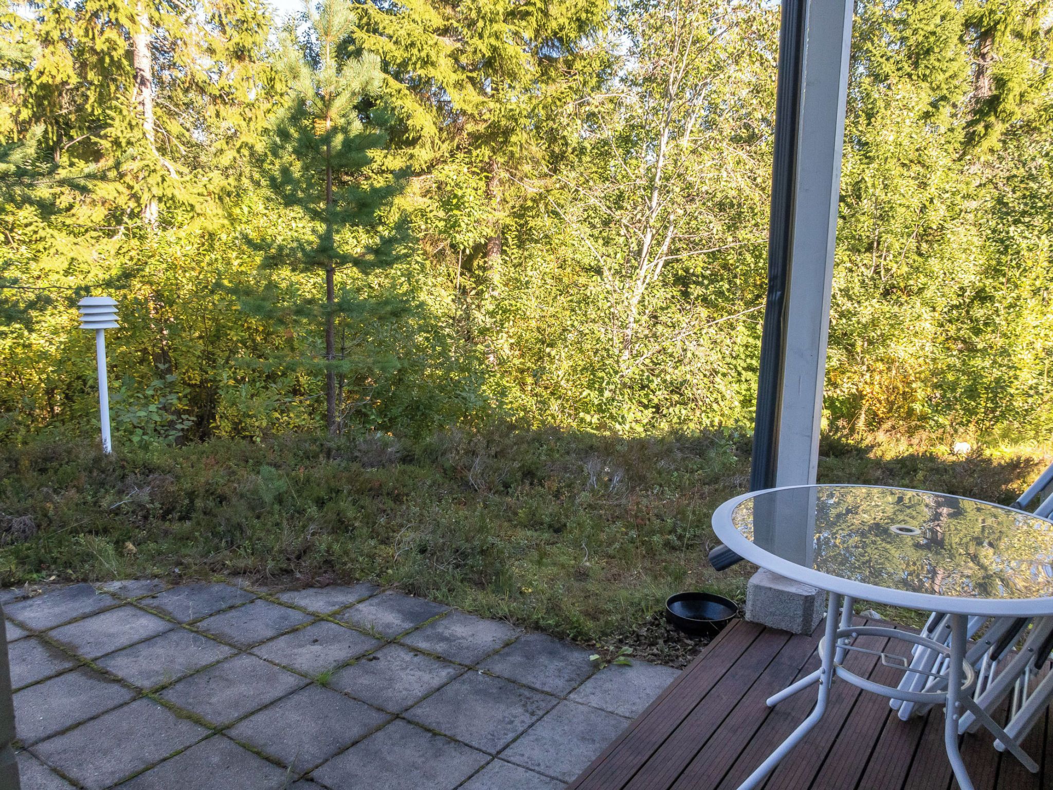 Un patio al aire libre con una mesa y sillas de vidrio, rodeado por una terraza de madera y un camino de piedra.