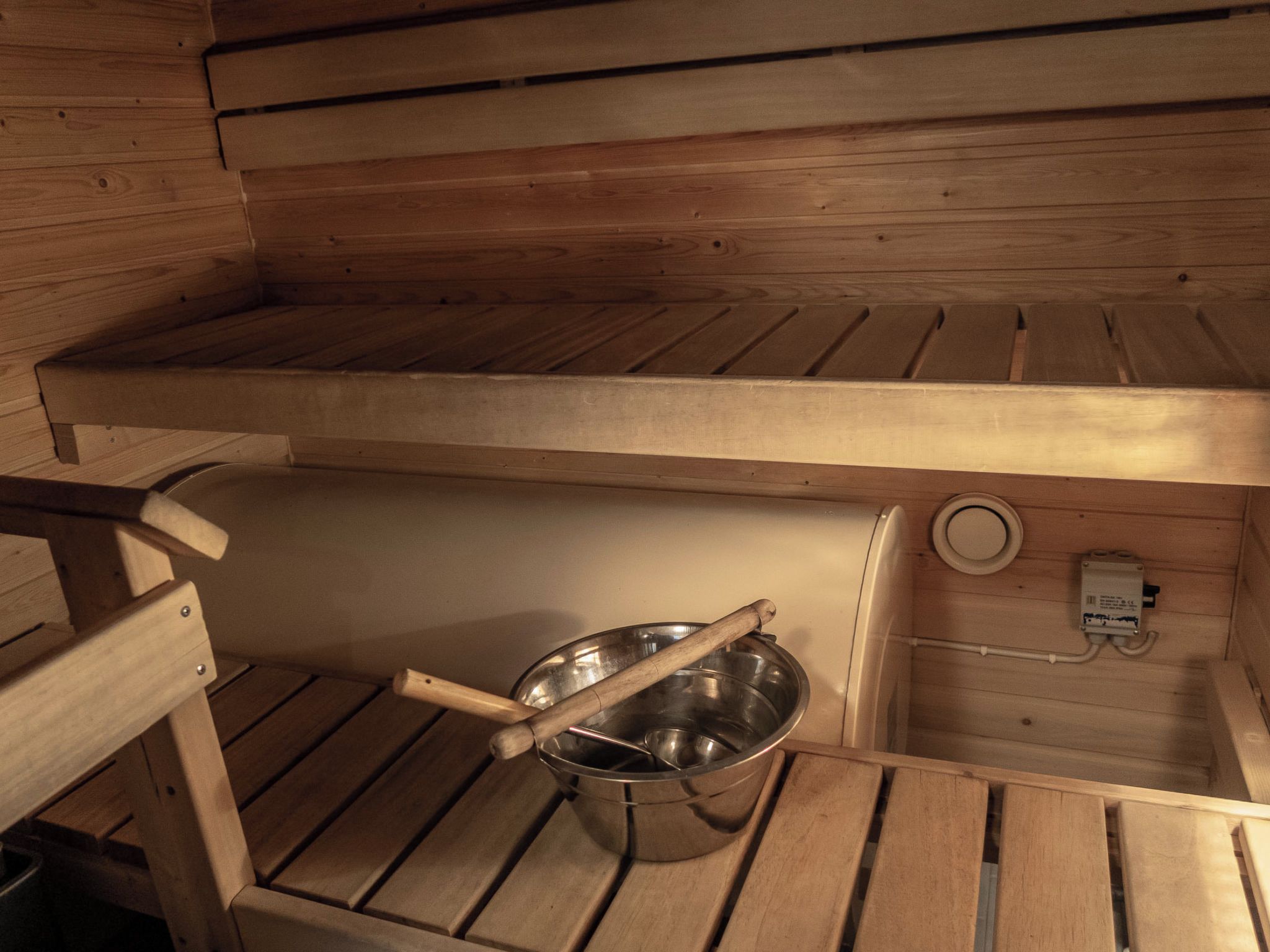 A sauna with wooden benches, a metal basin with a wooden handle, and a control unit on the wall.