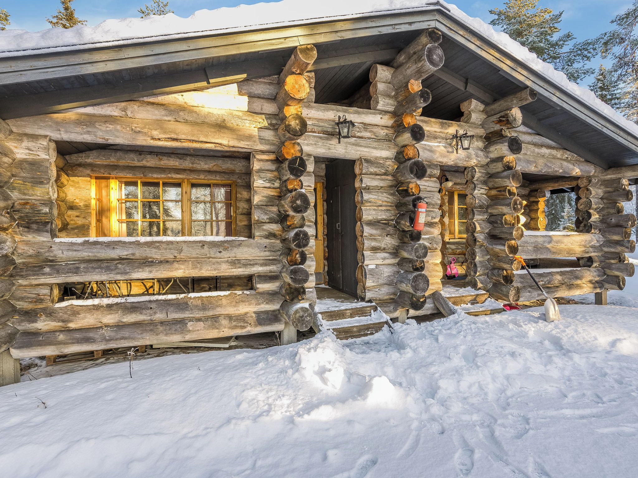 A log cabin with a snow-covered roof, large windows, and a fire extinguisher mounted on the exterior wall.