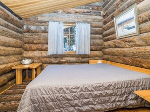 Quarto de cabana de madeira com uma cama, uma mesinha e uma janela com cortinas.