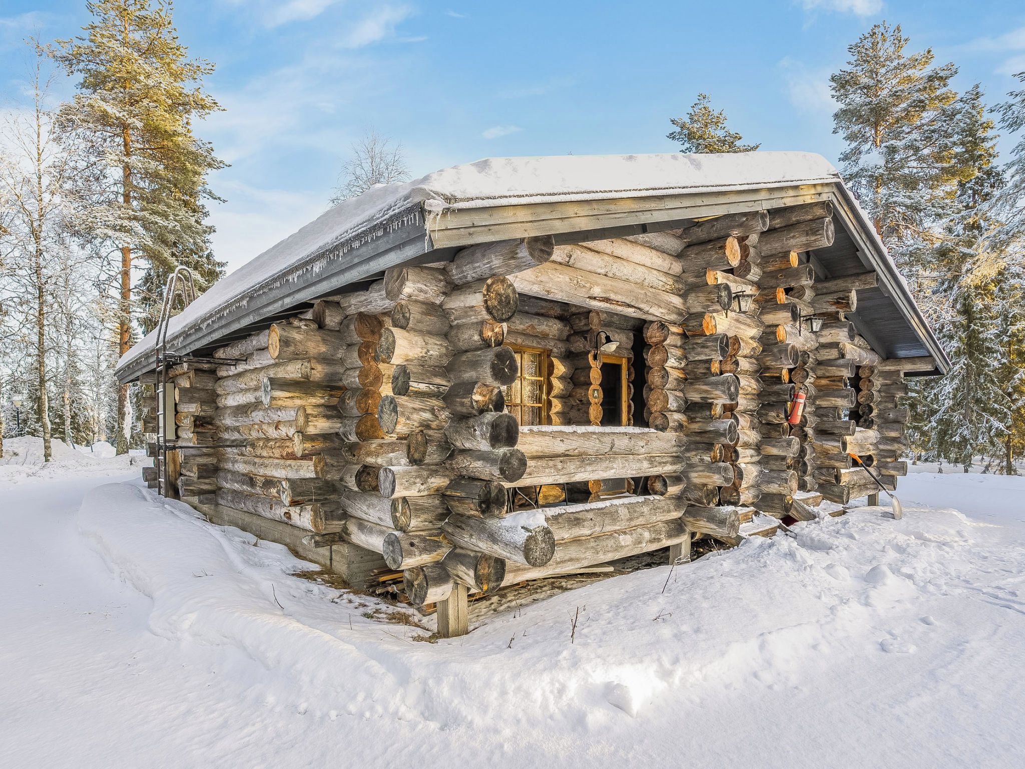 A log cabin with a snow-covered roof, surrounded by snow and trees.