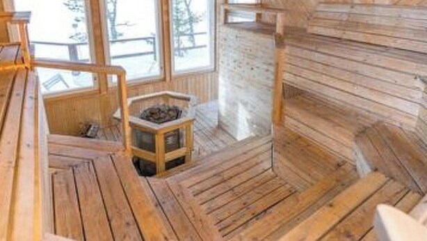 A wooden cabin interior with a central fireplace, wooden benches, and a window overlooking a snowy landscape. - Hästöskata a by Interhome (Kruunupyy)