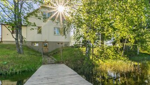 Une jetée en bois menant à une maison située au bord d’une étendue d’eau calme, le tout entouré d’une végétation luxuriante.
