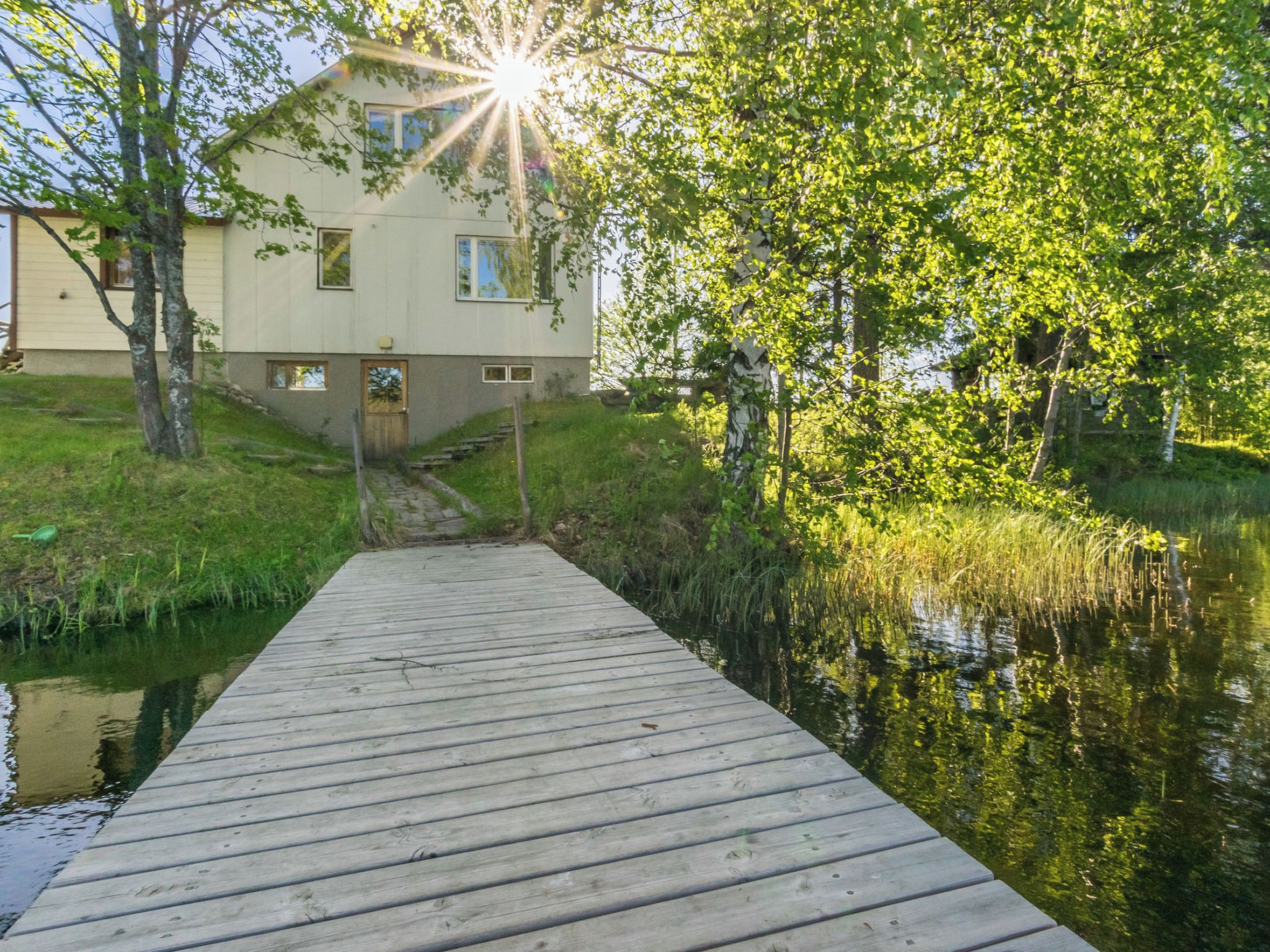A wooden dock leading to a house with a grassy area and trees around.