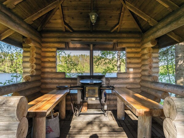 A wooden cabin with a central cooking area, benches, and a view of a forested area.