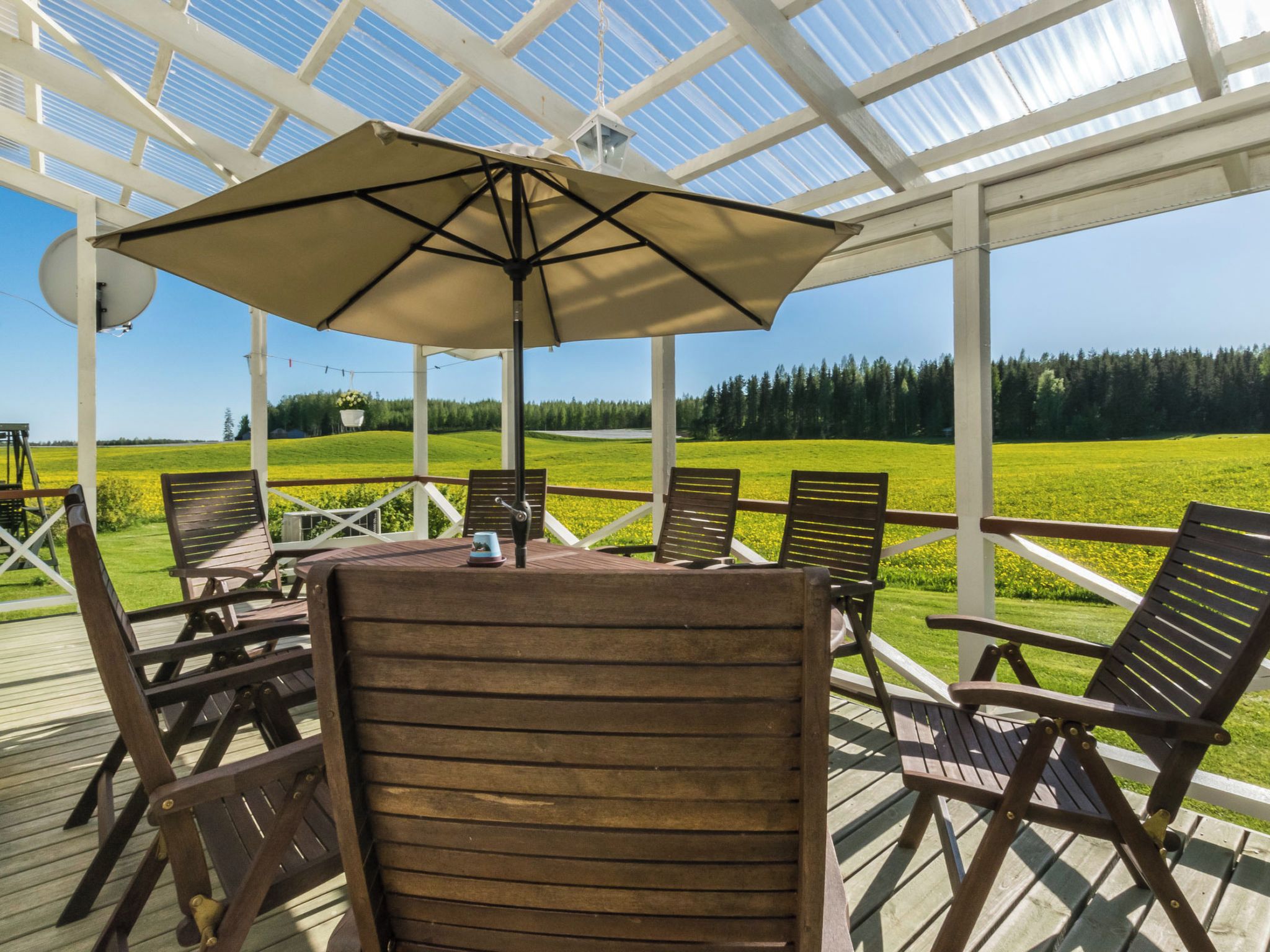 An outdoor patio with wooden chairs and a table, a large umbrella, and a view of a green field and trees.