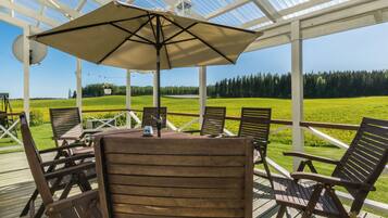 An outdoor patio with wooden chairs and a table, a large umbrella, and a view of a green field and trees.