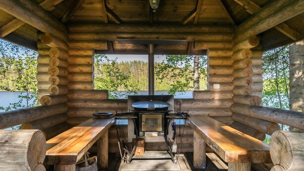 Une cabane en bois rustique, dotée d’une table centrale et de bancs, de grandes fenêtres et offrant une vue sur une forêt et un lac.