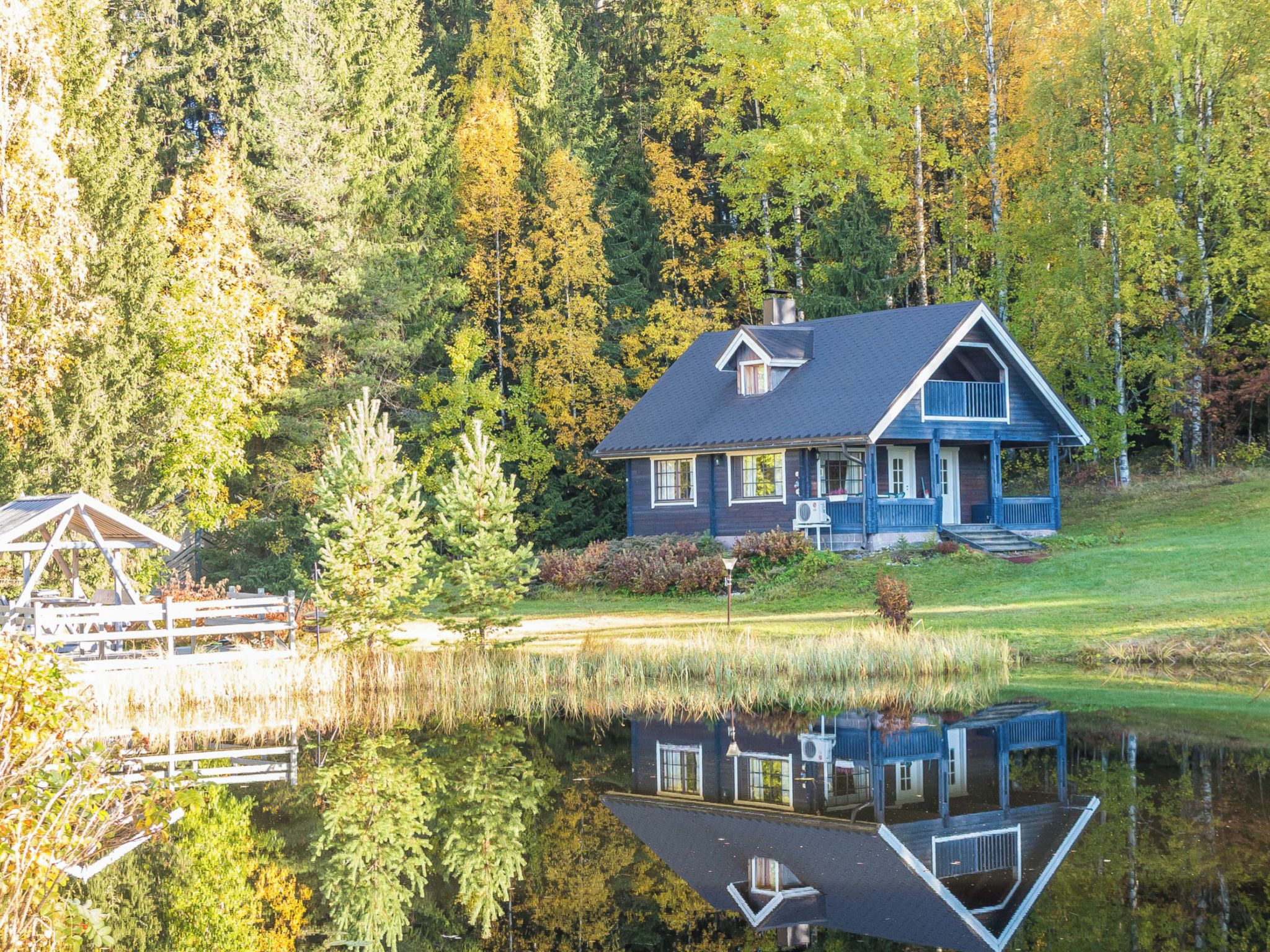 A house with a reflection in a calm lake, surrounded by trees and a wooden dock.