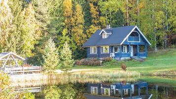 A house with a reflection in a calm lake, surrounded by trees and a wooden dock.