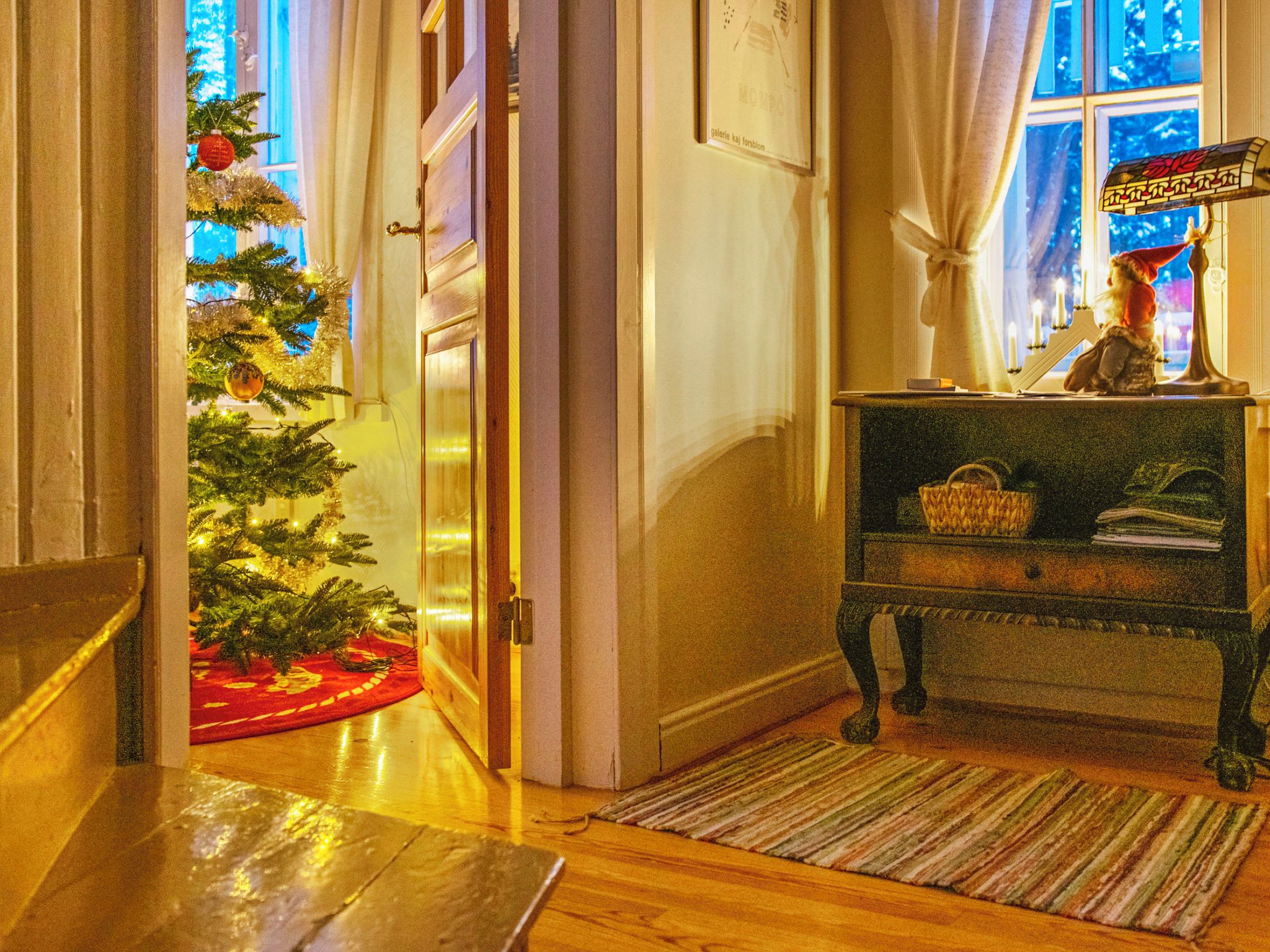 Un couloir décoré d’un sapin de Noël, un banc en bois avec un panier, et une bougie allumée posée sur une table.