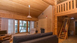 L’intérieur chaleureux d’un chalet avec un plafond en bois, une cheminée, un canapé, un téléviseur et un escalier menant à un étage supérieur.