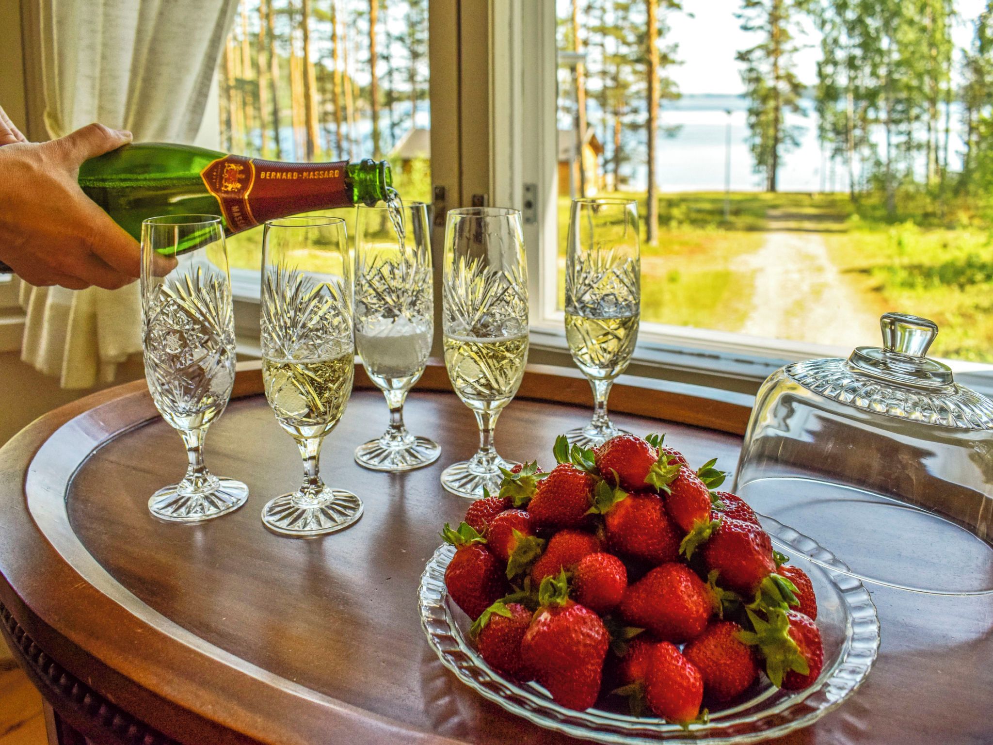 Ein Tisch mit Erdbeeren in einer Schale und einer Champagnerflasche, aus der Sekt in Gläser gefüllt wird.