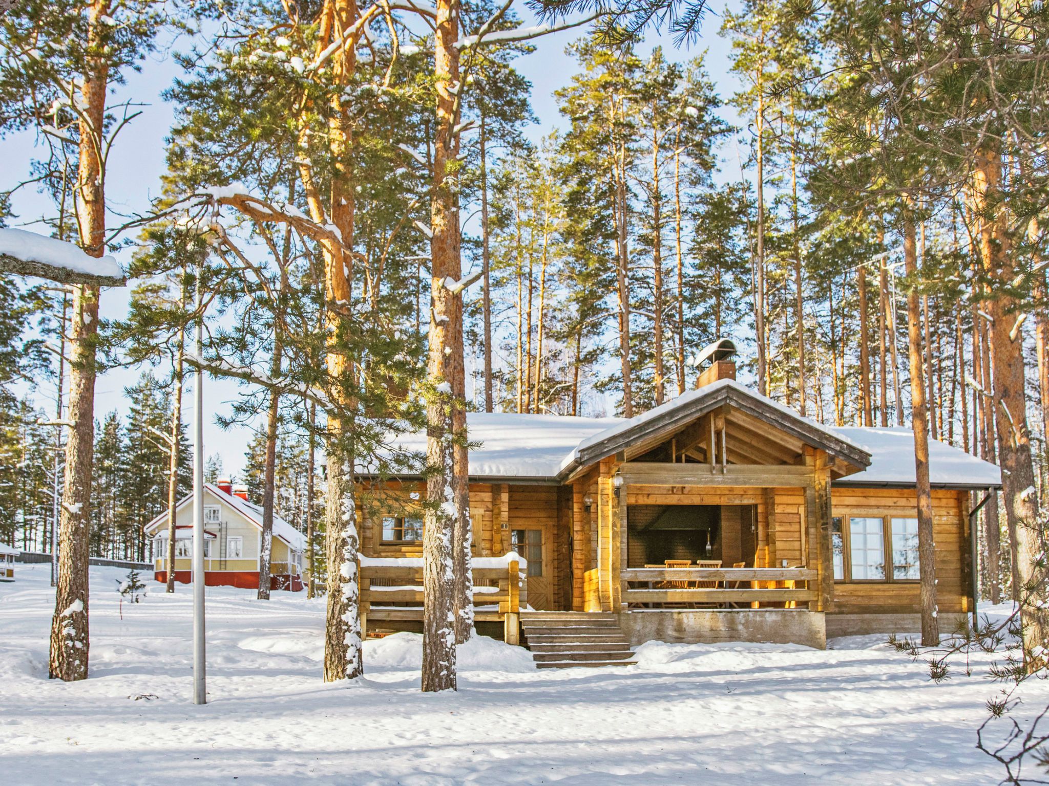 Puinen mökki, jossa on katettu kuisti ja takka, ja jonka ympärillä kohoaa korkeita mäntyjä lumisessa maisemassa.