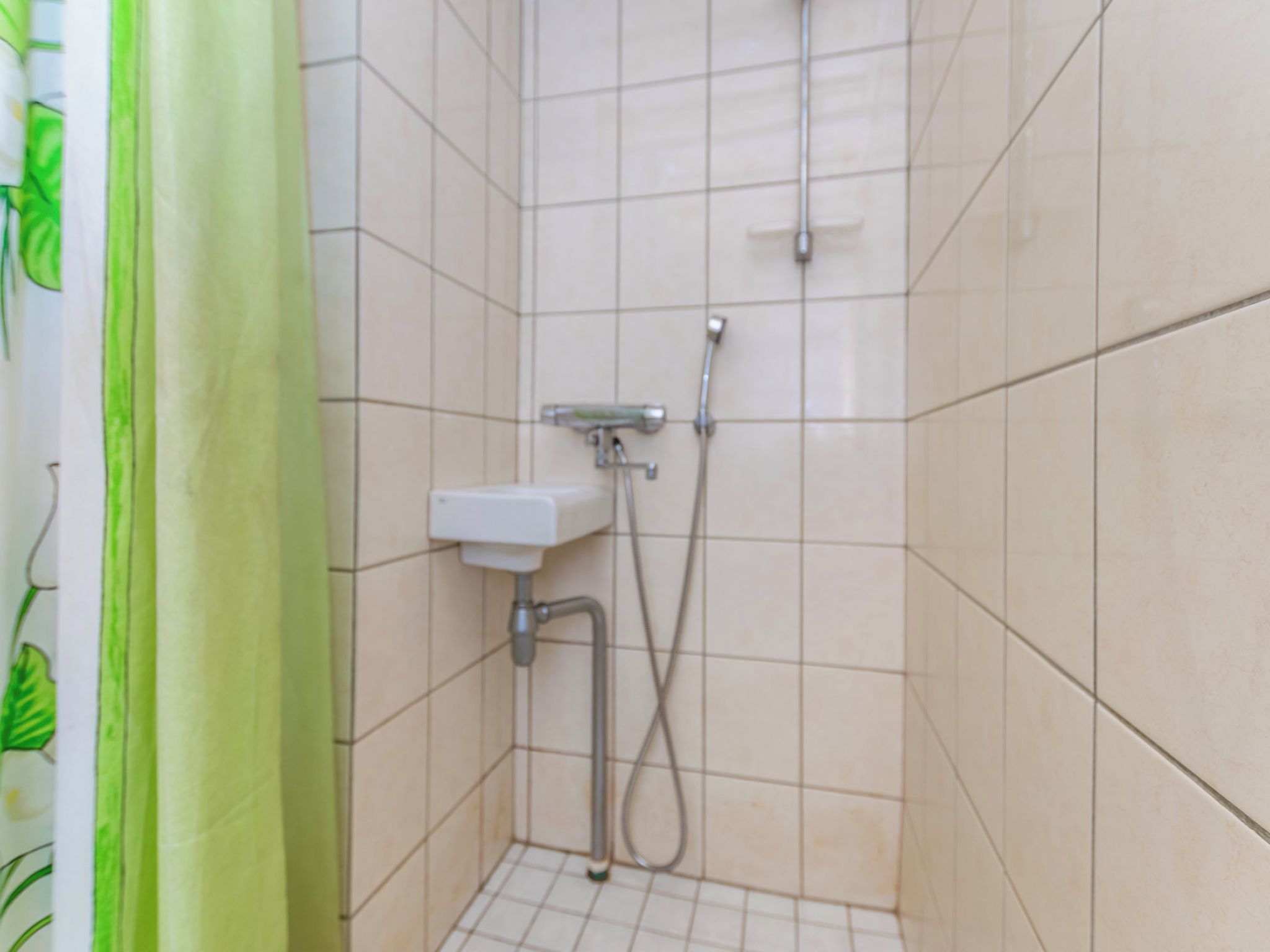 Eine kleine, geflieste Duschkabine mit einer Handbrause und einem am Wand montierten Wasserhahn.