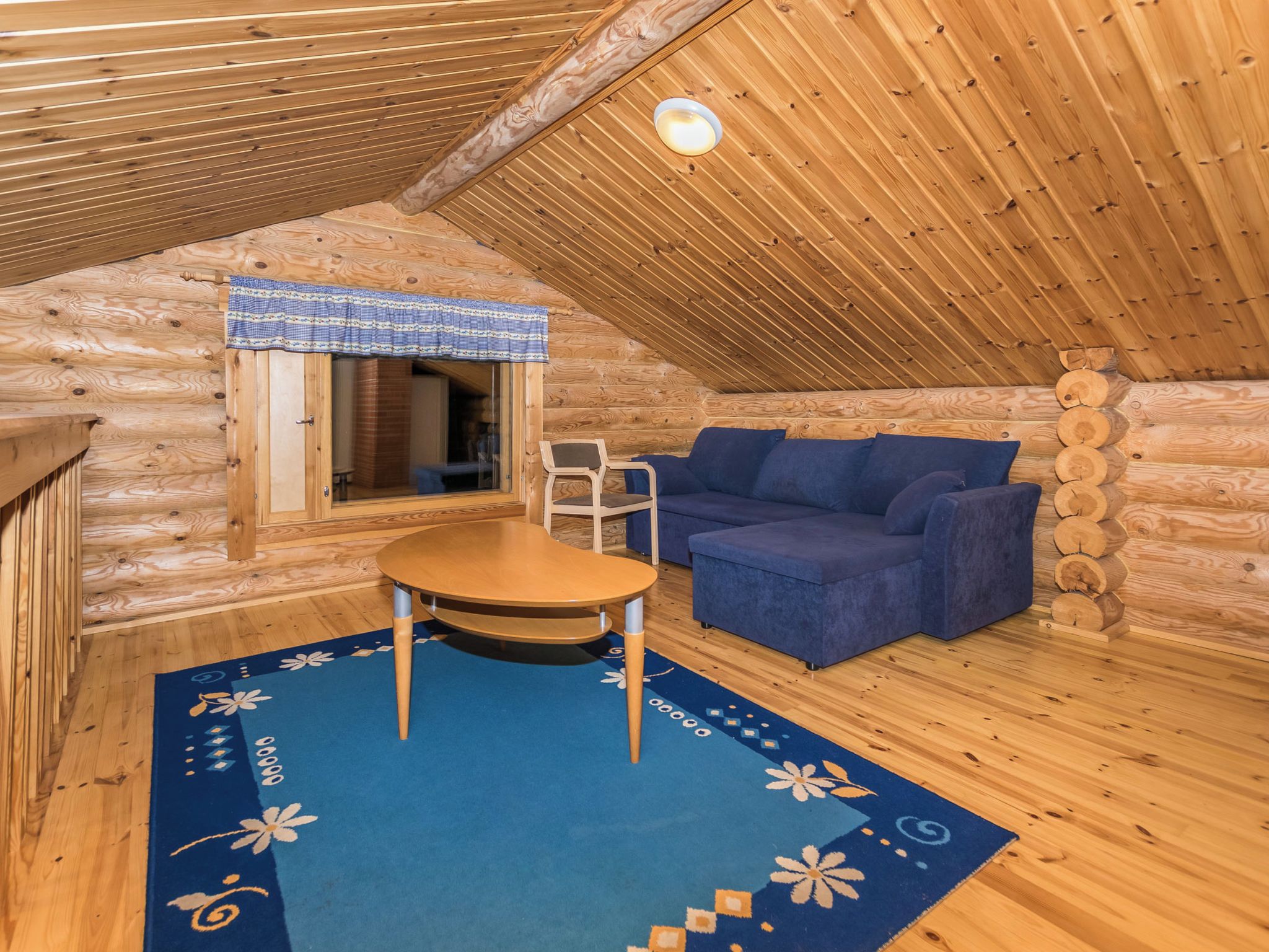 Interior acogedor de una cabaña de troncos con techo de madera, una mesa redonda, un sofá azul y una ventana con cortina a cuadros.