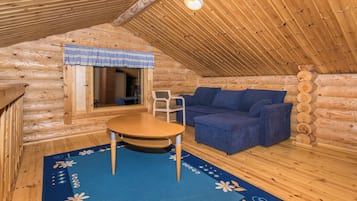 Interior acogedor de una cabaña de troncos con techo de madera, una mesa redonda, un sofá azul y una ventana con cortina a cuadros.