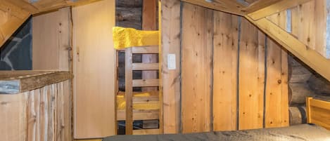 A loft bedroom with a sloped ceiling, wooden walls, a ladder leading to a lofted sleeping area, and a bed with a grey quilt.