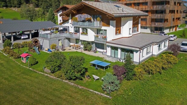 Un edificio moderno de varios pisos con balcones, una zona de estar al aire libre cubierta y montañas al fondo.