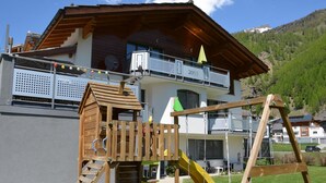 Un edificio de dos pisos con un parque infantil de madera, que incluye un tobogán y columpios, al frente.