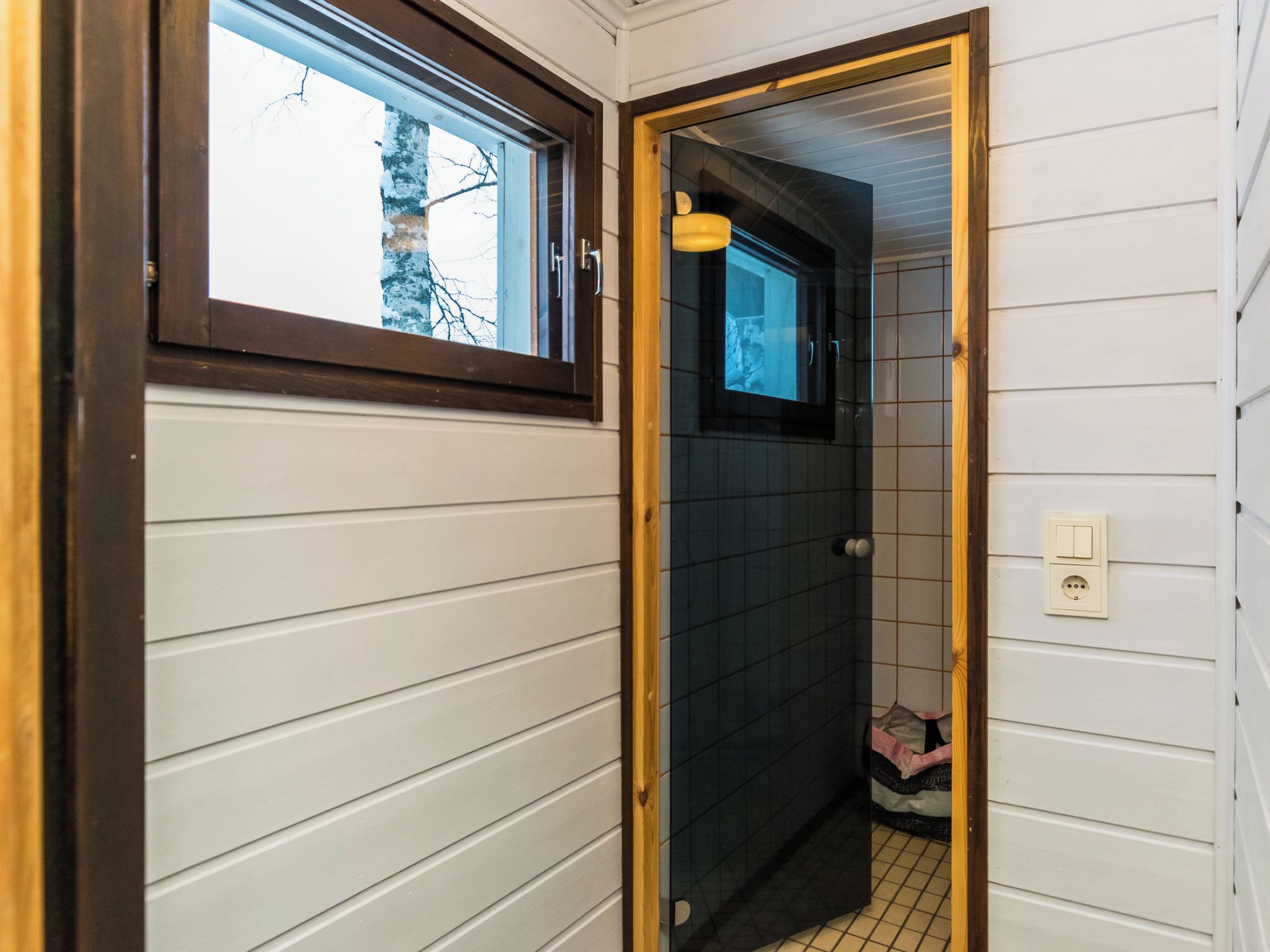 A narrow hallway with a window on the left and a door leading to a bathroom with dark tiles.