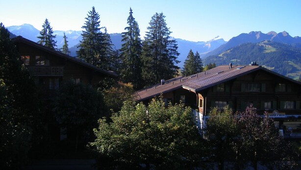 A mountain lodge with multiple wooden buildings, surrounded by tall evergreen trees and a backdrop of snow-capped mountains.