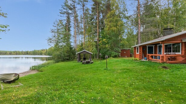 Une cabane dotée d’une terrasse et d’un petit abri, entourée d’une zone herbeuse et de grands arbres.